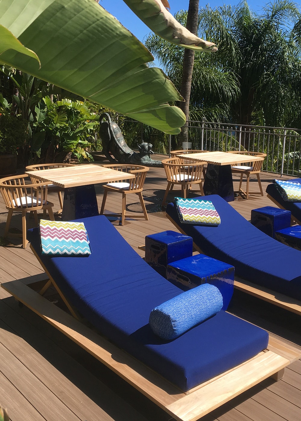 Terrasse ensoleillée avec chaises longues bleues, tables en bois et végétation luxuriante, idéale pour se détendre sous un ciel bleu sans nuages.