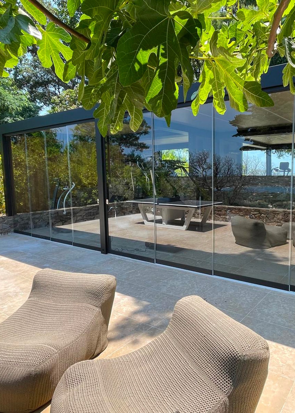 Terrasse moderne avec fauteuils confortables, grande baie vitrée et feuilles d'arbre. L'intérieur reflète le jardin environnant sous une lumière naturelle apaisante.