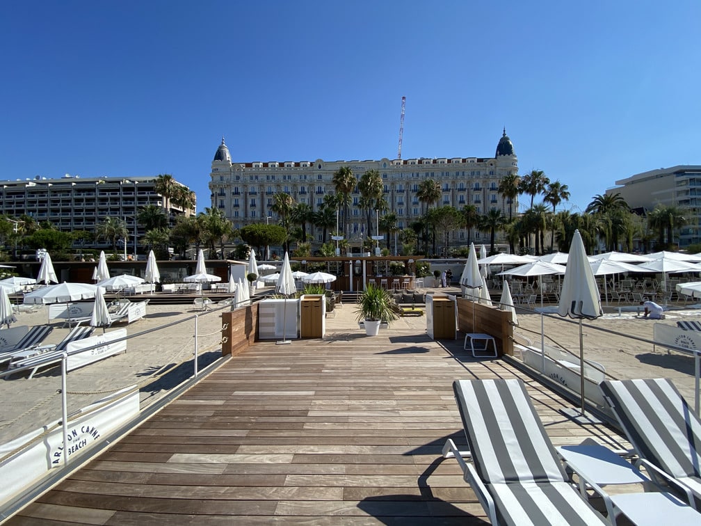 Plage ensoleillée avec chaises longues, parasols et le célèbre hôtel Carlton en arrière-plan, à Cannes. Palmiers et ciel bleu complètent la scène estivale.