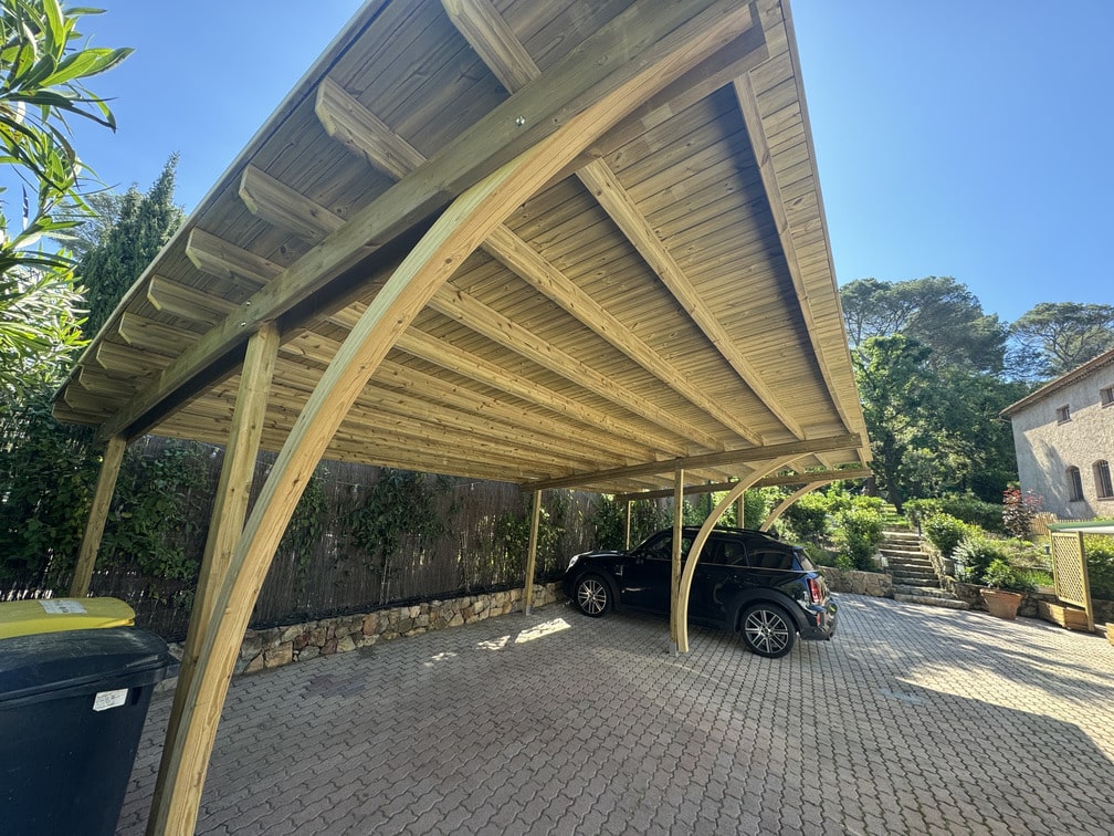 Voiture garée sous un carport en bois, entourée de verdure. Pavage et bâtiment en pierre à proximité. Ciel clair et arbres en arrière-plan.