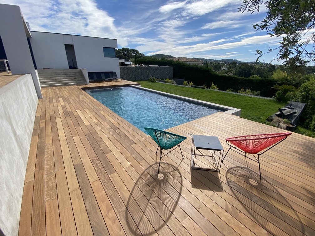 Terrasse en bois avec piscine, deux chaises colorées et table. Vue sur un jardin et collines sous un ciel partiellement nuageux.