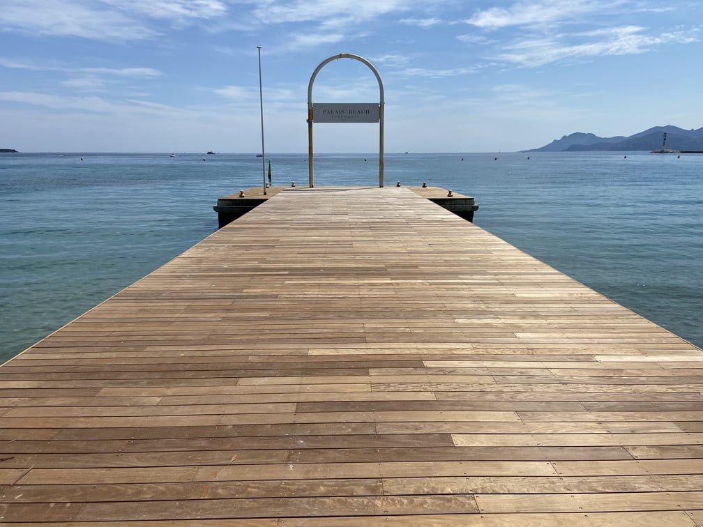 Une jetée en bois mène à l'océan sous un ciel bleu. Panneau indiquant "Plage". Montagnes visibles à l'horizon. Atmosphère paisible.