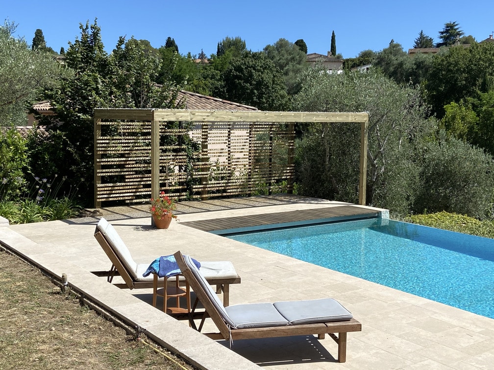 Transats près d'une piscine à débordement, pergola minimaliste en bois entourée de verdure. Ciel bleu, ambiance tranquille et estivale. Personne visible sur les chaises longues.