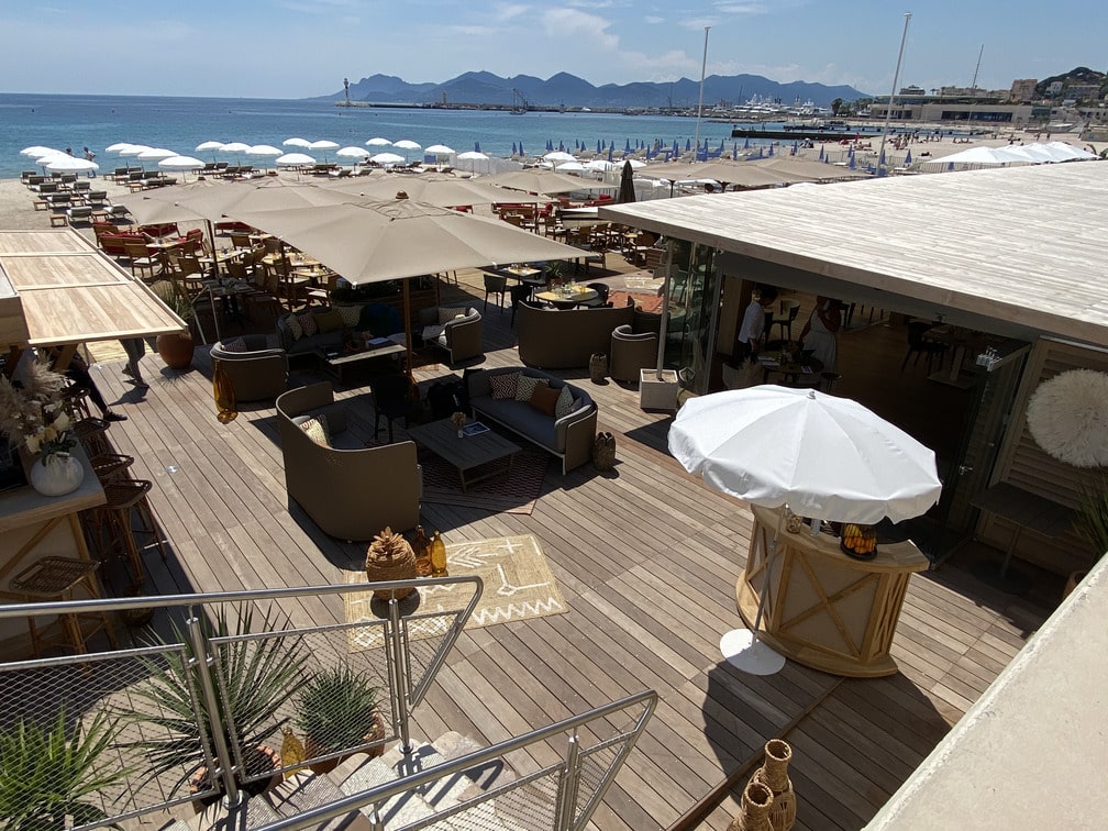 Une terrasse en ipé sur la plage de Cannes, avec parasols et chaises longues. Vue sur la mer et les montagnes au loin.