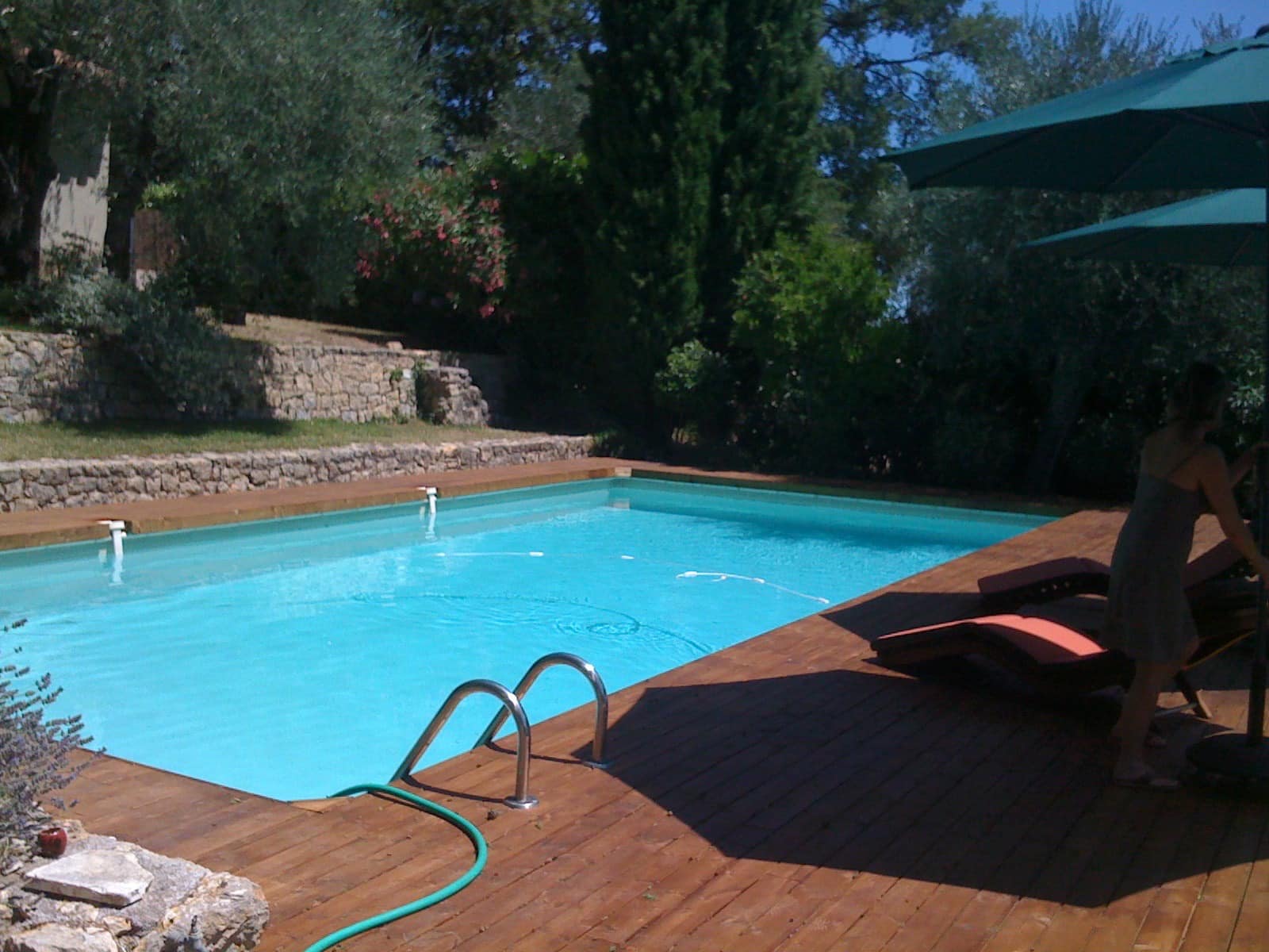 Une piscine entourée de bois et de verdure. Une personne se tient à côté, profitant de l'ombre de parasols verts.