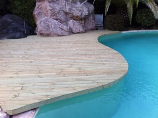 Dalle en bois entourant une piscine turquoise, avec des rochers et des plantes en arrière-plan. Aucune personne n'est visible dans l'image.