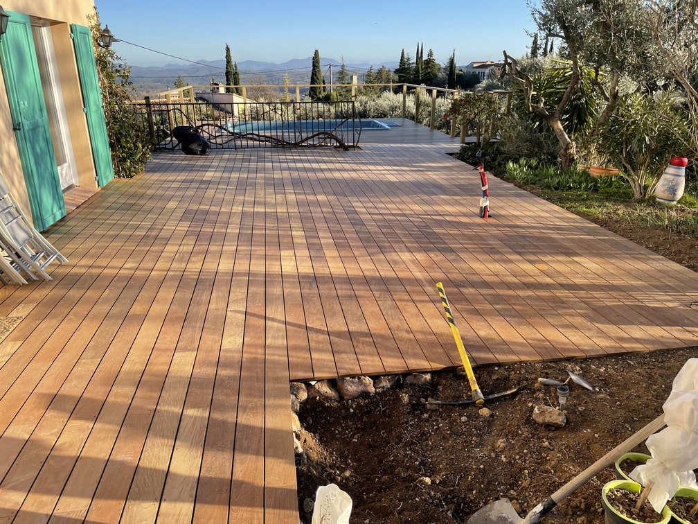 Terrasse en ipé en construction près d'une piscine, avec vue sur la campagne et montagnes. Végétation dense autour, ciel dégagé, outils visibles.