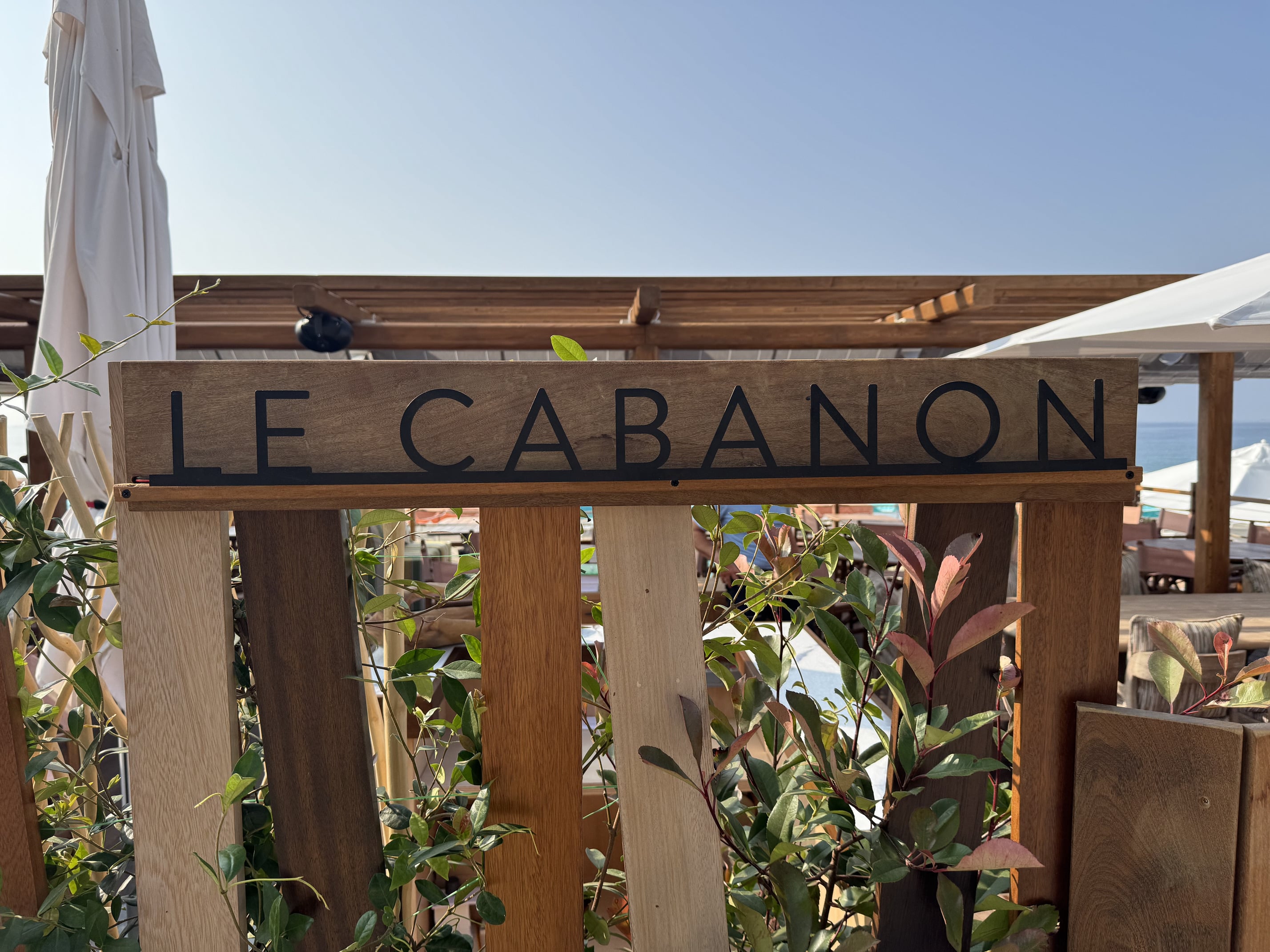 Un panneau en bois avec l'inscription "Le Cabanon" entouré de plantes, surplombant la mer sous un ciel dégagé et ensoleillé.