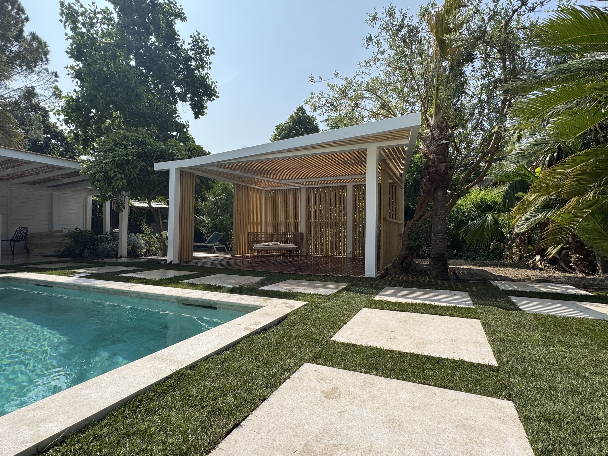 Un jardin avec piscine, pergola moderne en bois, entouré de palmiers et d'arbres verts, ciel dégagé, atmosphère paisible et ensoleillée.